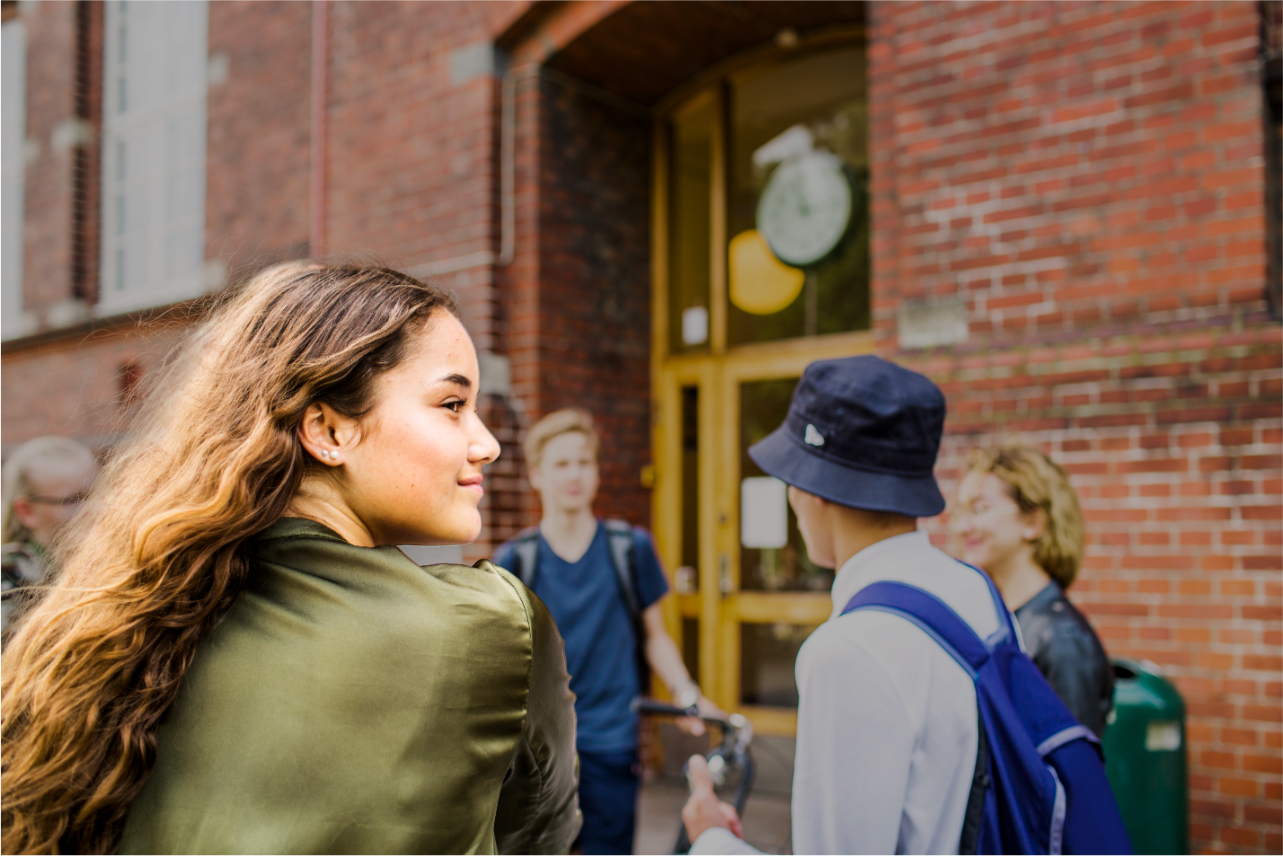 A group of students gather outside a brick school building, smiling and talking