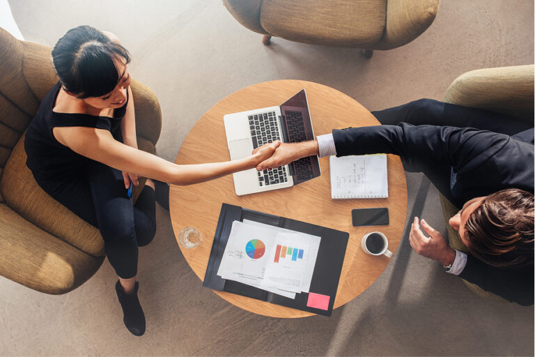Two professionals shake hands at a round table with charts and a laptop