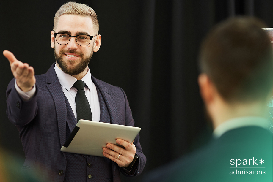 A man in a suit gestures while holding a tablet and speaking to an audience, highlighting leadership skills for business majors