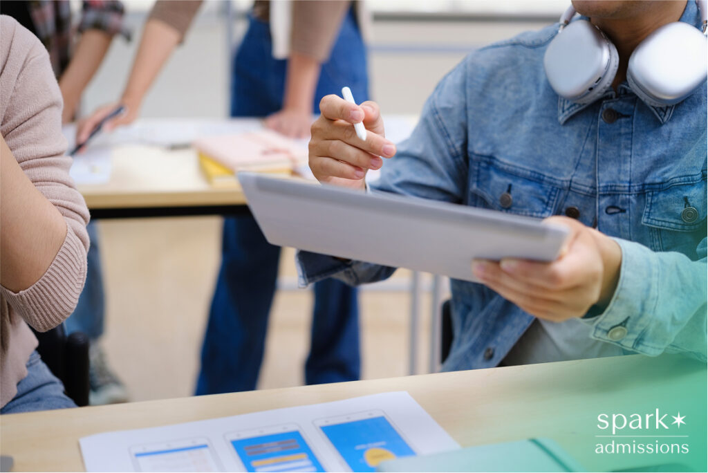 A student holds a tablet and stylus while discussing a digital project at a classroom table