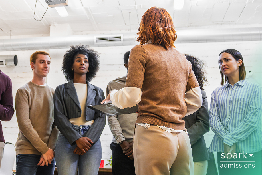 A woman holding a clipboard speaking to a diverse group of students, symbolizing how colleges may verify extracurricular activities through interviews or evaluations