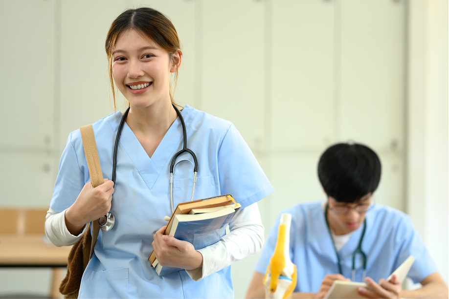 A smiling nursing student holds books and a stethoscope in a classroom