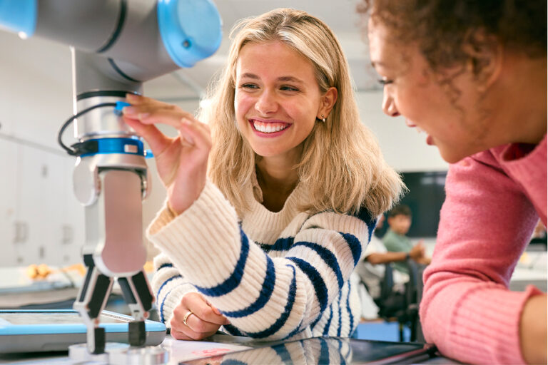 Two female students smile while working with a robotic arm in a classroom