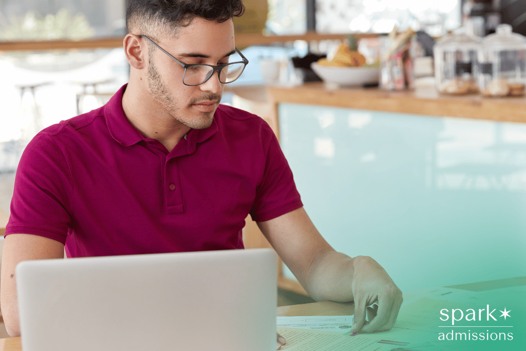 Man wearing glasses writing a transfer essay on laptop while reviewing notes on paper