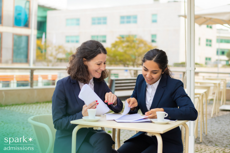 Two female business college students smiling while reviewing papers together at a café table